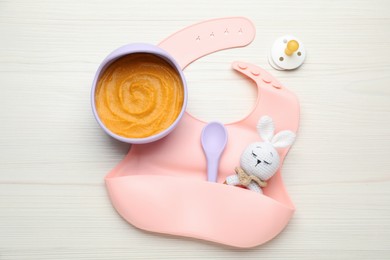 Silicone baby bib, toy and plastic dishware with healthy food on white wooden table, flat lay