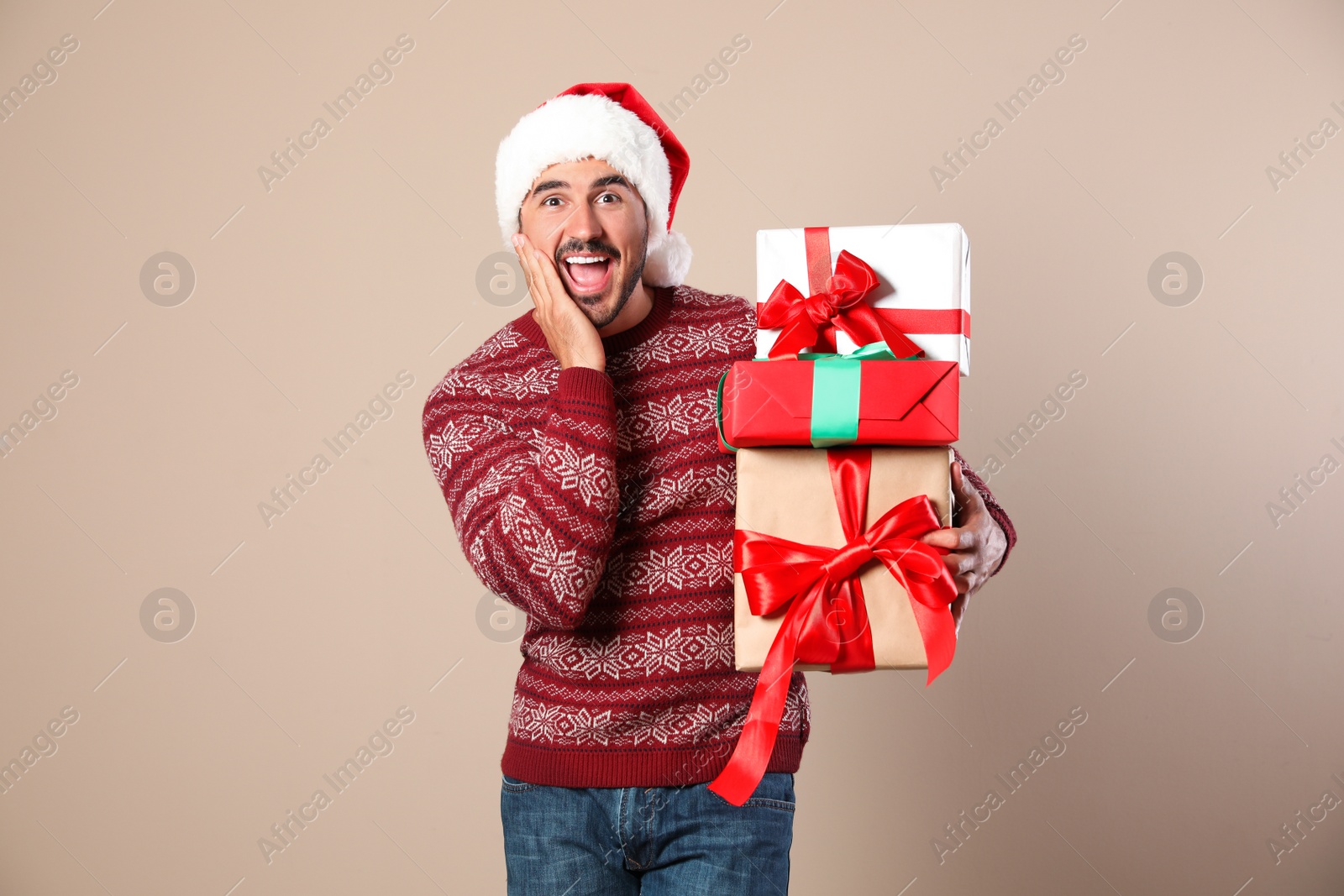 Photo of Happy man in Christmas sweater and Santa hat holding gift boxes on beige background