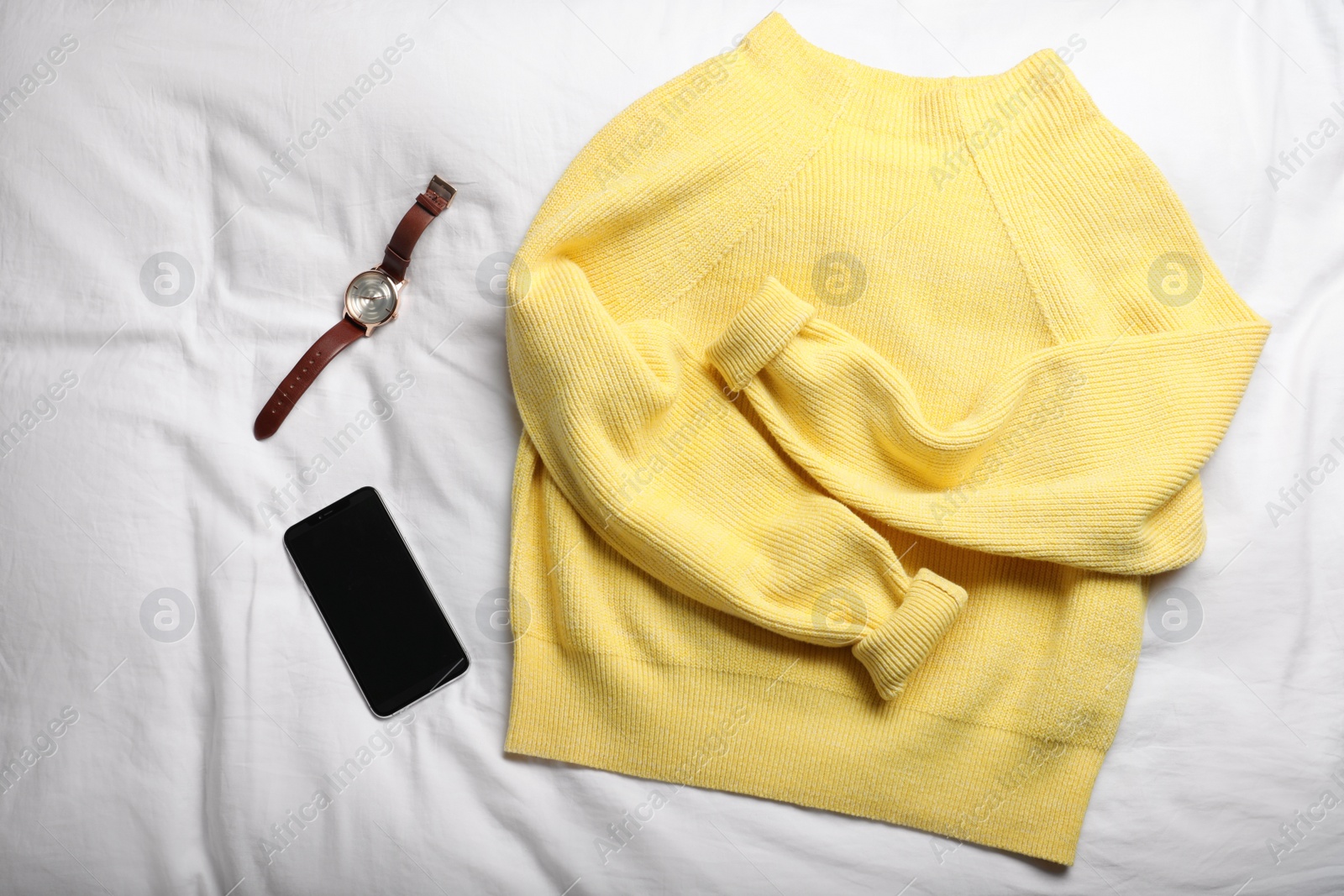 Photo of Yellow warm sweater with smartphone and wristwatch on bedsheet, flat lay