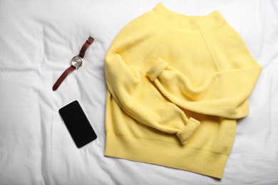 Photo of Yellow warm sweater with smartphone and wristwatch on bedsheet, flat lay