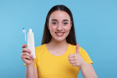 Photo of Happy young woman with plastic toothbrush and tube of toothpaste showing thumb up on light blue background