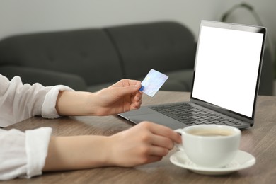 Photo of Woman with credit card and cup of coffee near laptop at wooden table indoors, closeup. Online shopping