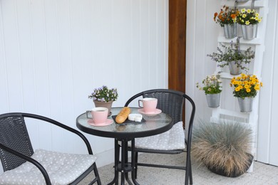 Photo of Cups of coffee, potted plant, bread and cheese on glass table. Relaxing place at outdoor terrace