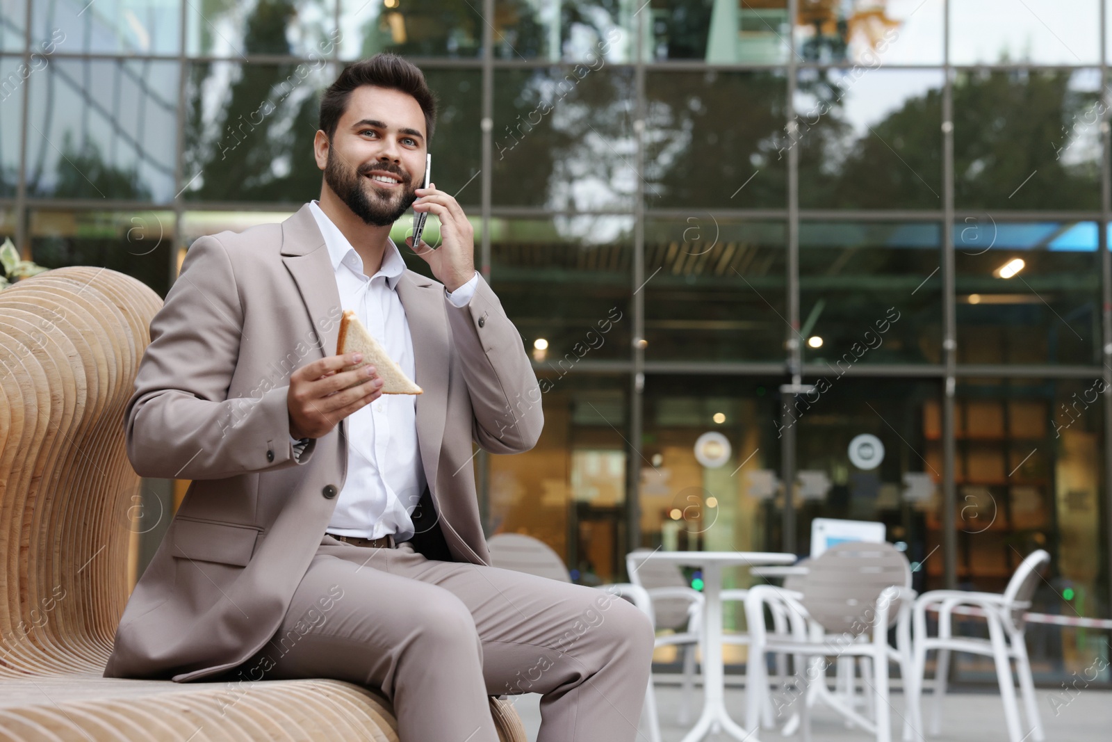 Photo of Lunch time. Young businessman with sandwich talking on smartphone on bench outdoors