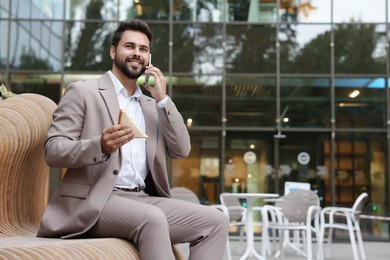 Photo of Lunch time. Young businessman with sandwich talking on smartphone on bench outdoors