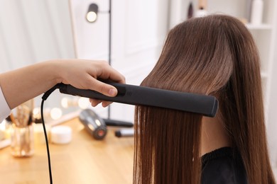 Photo of Hairdresser straightening woman's hair with flat iron in salon, closeup