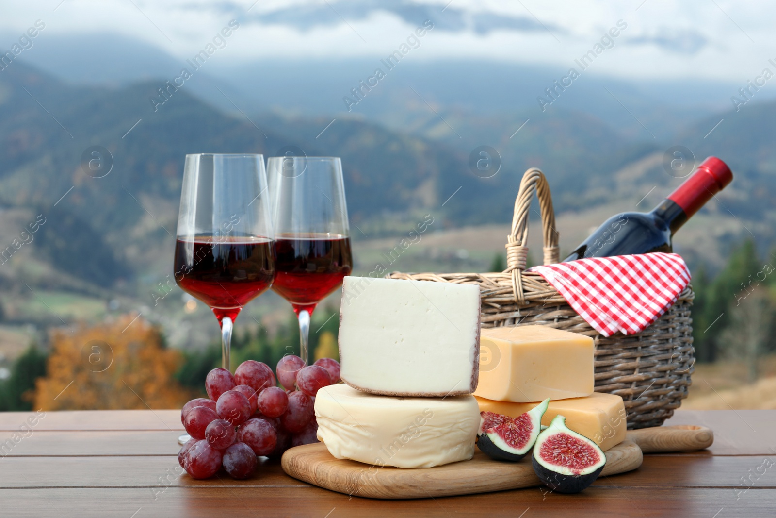 Photo of Different types of delicious cheeses, snacks and wine on wooden table against mountain landscape