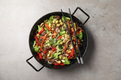 Photo of Stir-fry. Tasty noodles with meat in wok and chopsticks on grey textured table, top view