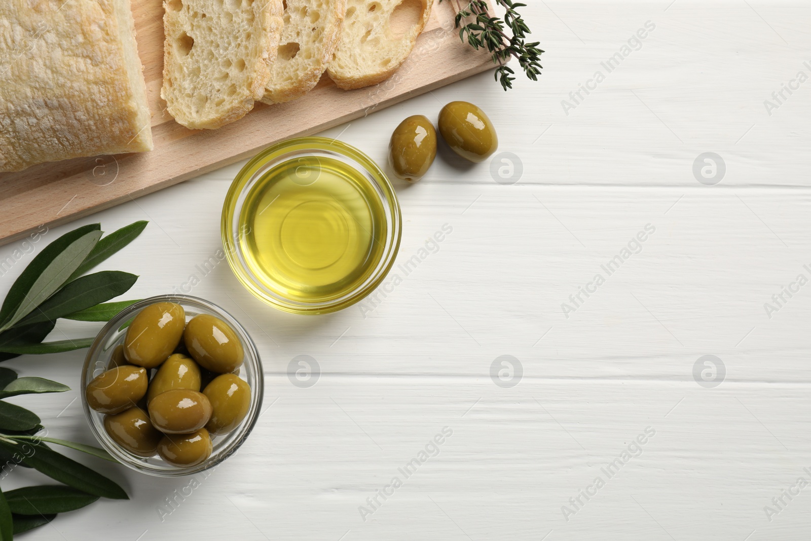 Photo of Bowl of cooking oil, ripe olives and bread on white wooden table, flat lay. Space for text