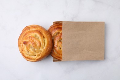 Photo of Paper bag with delicious rolls on white marble table, top view. Sweet buns