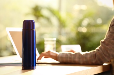 Woman working at table in modern office, focus on blue thermos bottle