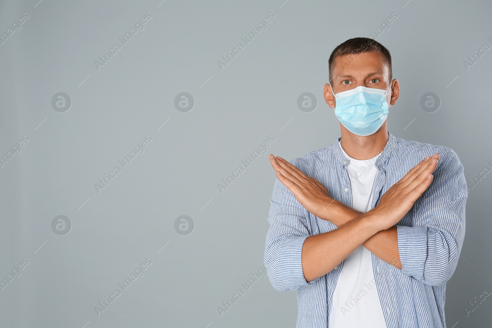 Photo of Man in protective mask showing stop gesture on grey background, space for text. Prevent spreading of coronavirus