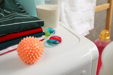 Photo of Orange dryer ball near stacked clean clothes and detergents on washing machine, closeup. Space for text