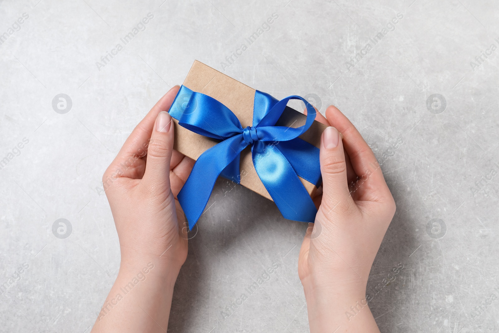 Photo of Woman holding gift box with blue bow at light grey table, top view