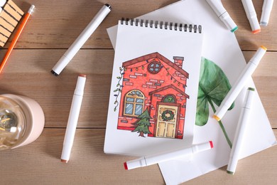 Photo of Sketchbook with beautiful drawing of house and felt tip pens on wooden table, flat lay