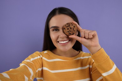 Photo of Young woman with chocolate chip cookie taking selfie on purple background