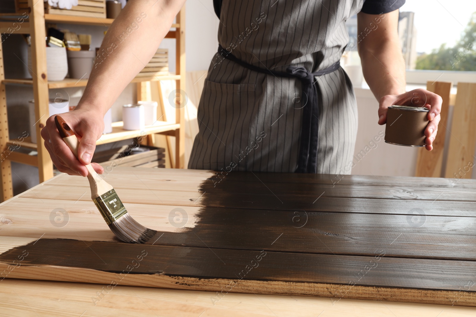 Photo of Man with brush and can applying wood stain onto wooden surface indoors, closeup