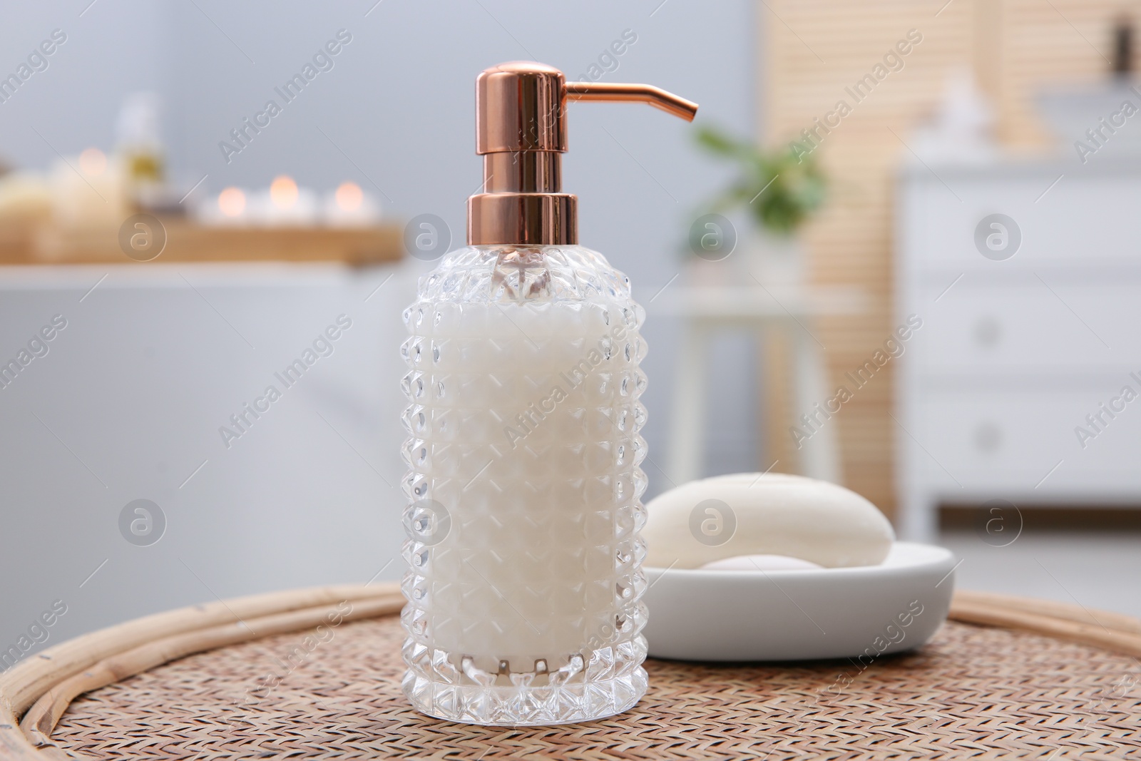 Photo of Stylish glass dispenser and soap bar on wicker stool in bathroom