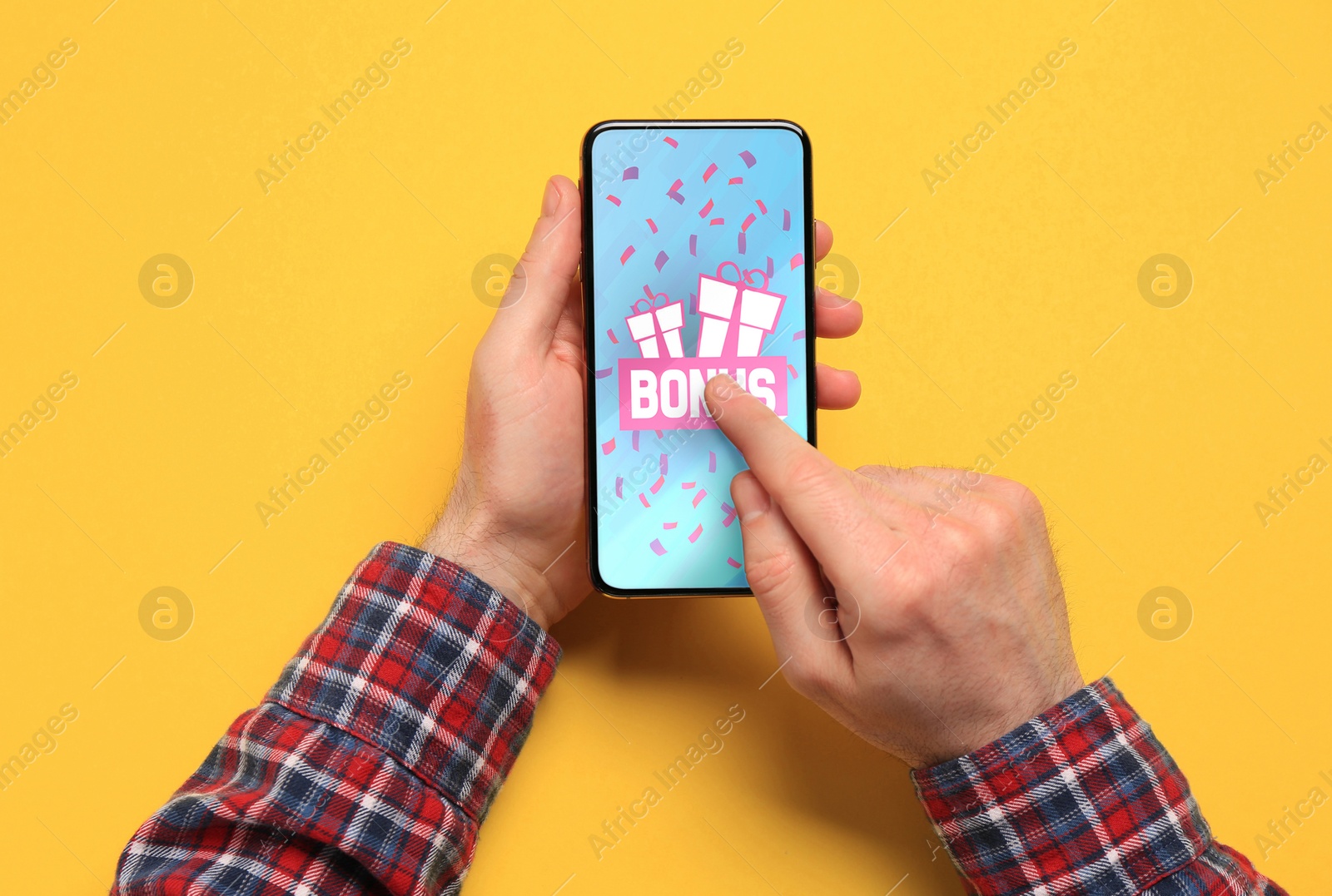 Image of Bonus gaining. Man using smartphone on yellow background, top view. Illustration of gift boxes, word and falling confetti on device screen
