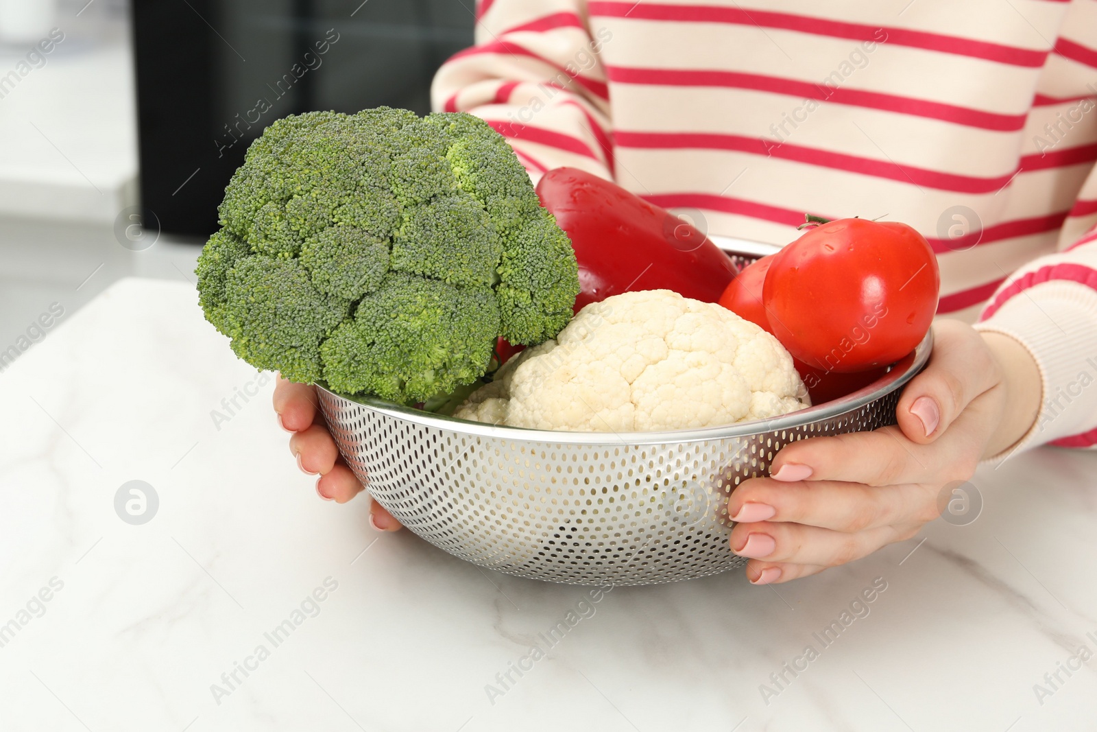 Photo of Woman holding colander with fresh vegetables at white marble table in kitchen, closeup