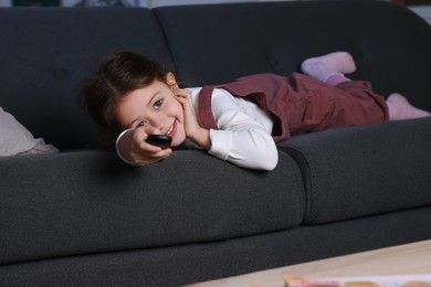 Photo of Cute little girl changing TV channels with remote control on sofa at home