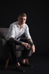 Young man with tattoos and glass of whiskey sitting in armchair on black background