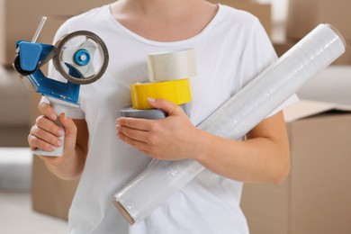 Photo of Woman with handheld dispenser, rolls of adhesive tape and plastic stretch wrap indoors, closeup