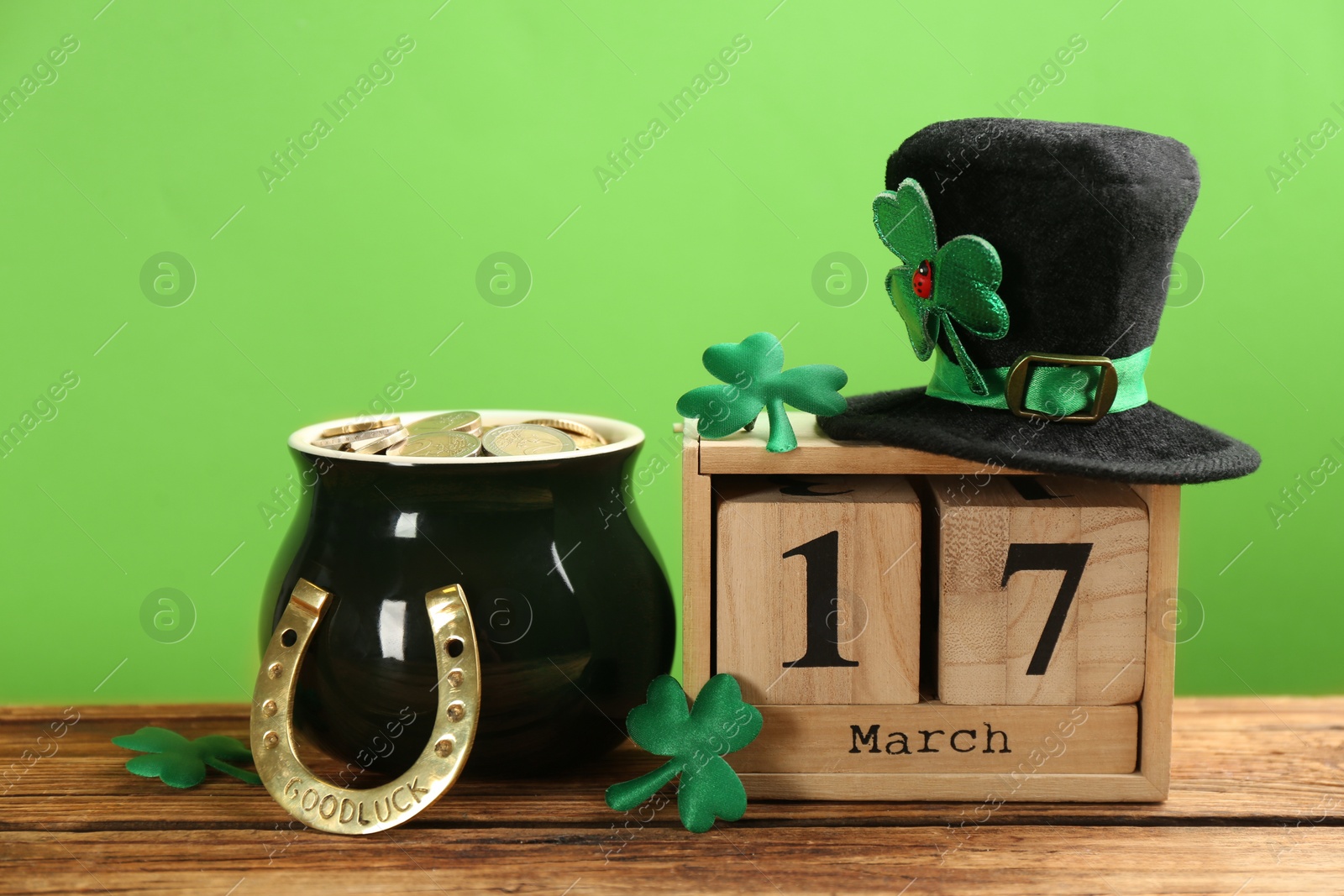 Photo of Composition with leprechaun hat, block calendar and pot of gold on wooden table against green background. St Patrick's Day celebration