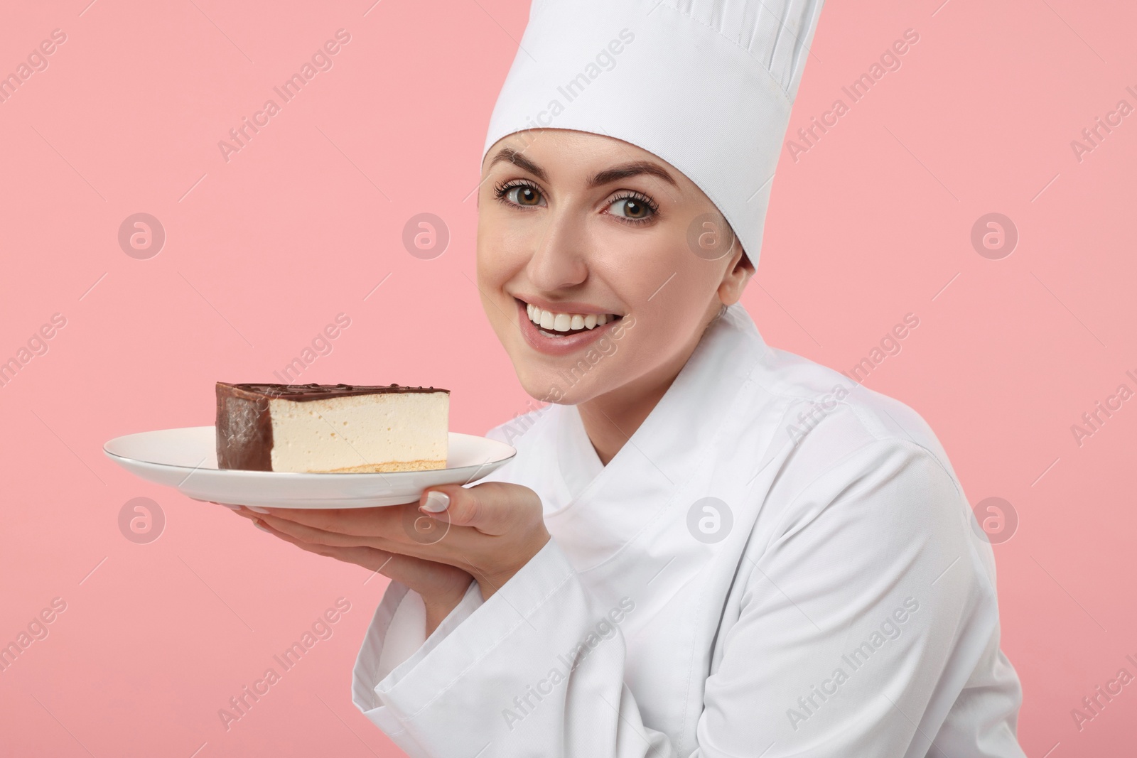 Photo of Happy professional confectioner in uniform holding delicious cheesecake on pink background