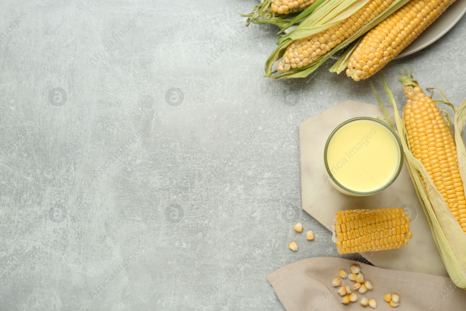 Photo of Tasty fresh corn milk in glass and cobs on light grey table, flat lay. Space for text