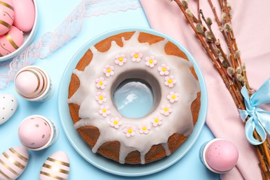 Photo of Delicious Easter cake decorated with sprinkles near painted eggs and willow branches on light blue background, flat lay