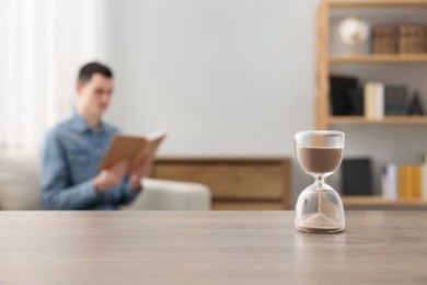 Photo of Hourglass with flowing sand on desk. Man reading book in room, selective focus