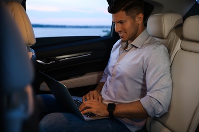 Photo of Handsome man working with laptop on backseat of modern car