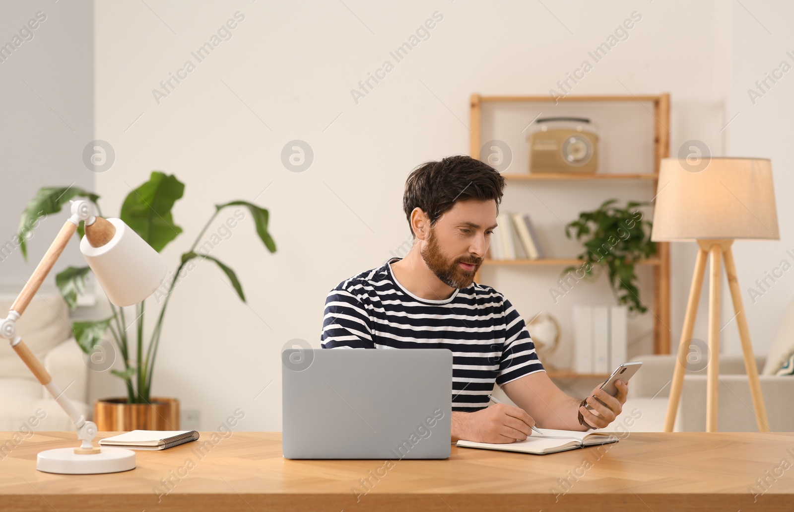 Photo of Home workplace. Man using smartphone and taking notes at wooden desk in room