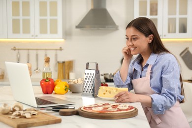 Happy woman making pizza while watching online cooking course via laptop in kitchen