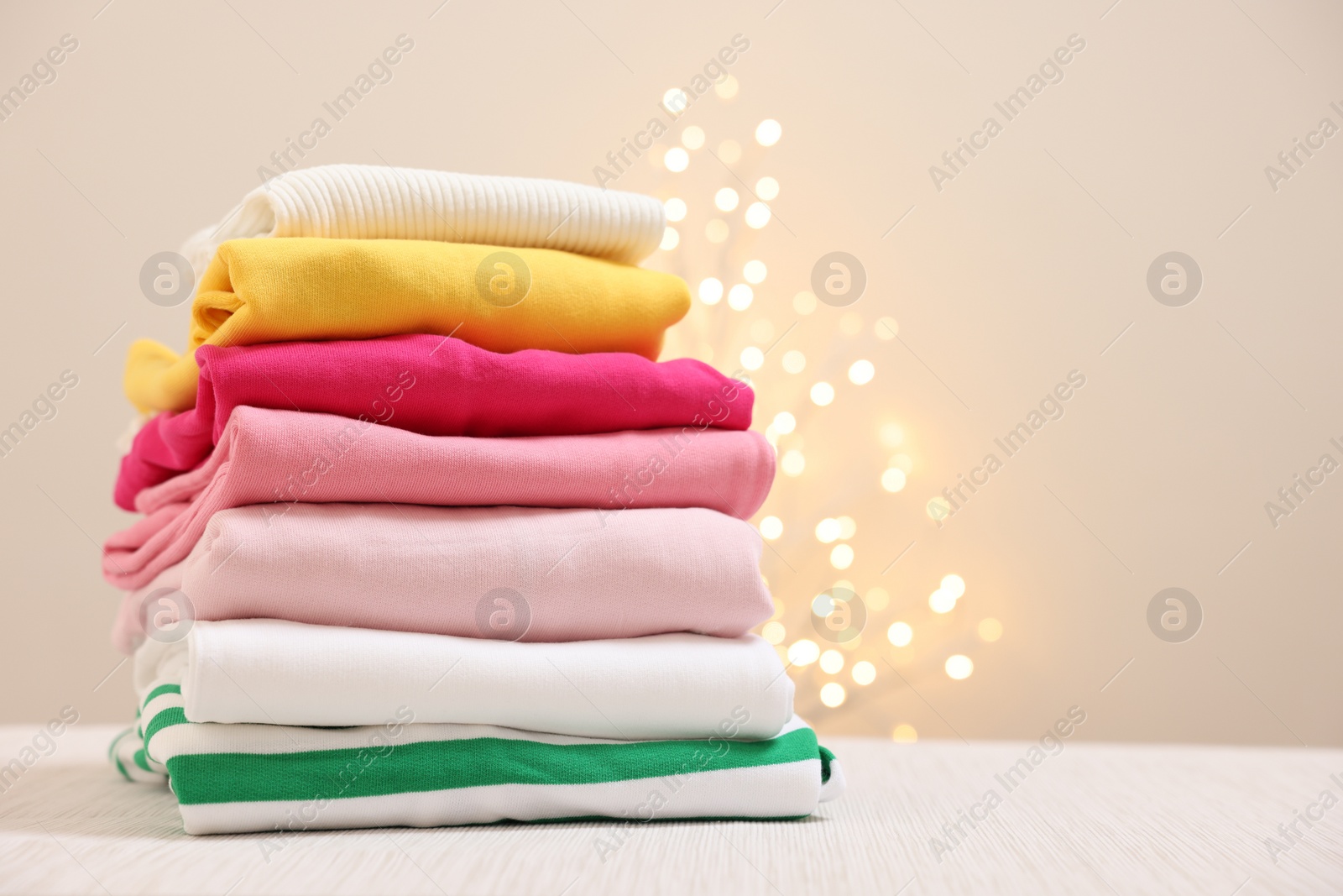 Stack of folded clothes on wooden table against beige background with blurred lights, space for text Photo of Stack of folded clothes on wooden table against beige background with blurred lights, space for text