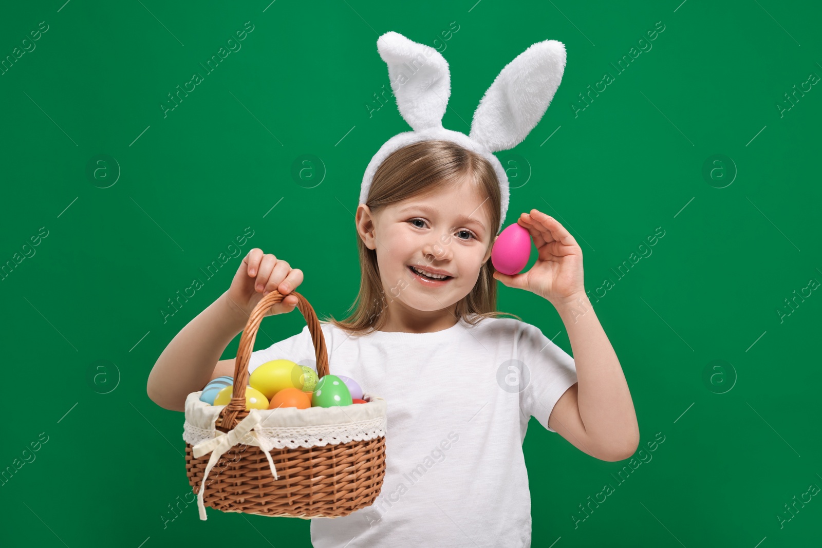 Photo of Easter celebration. Cute girl with bunny ears holding basket of painted eggs on green background