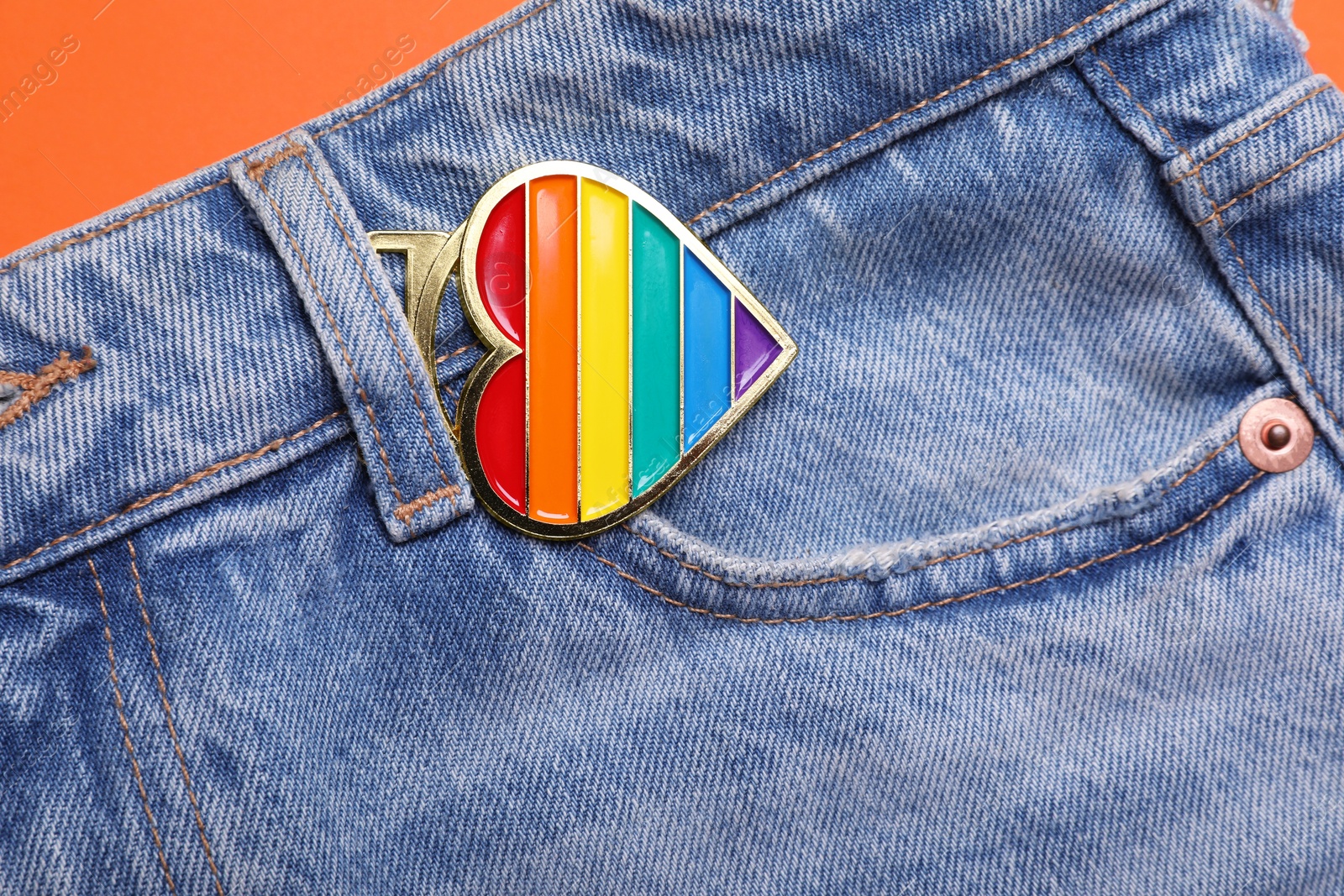 Photo of Jeans with rainbow heart shaped pendant on orange background, top view. LGBT pride