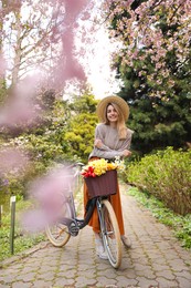 Beautiful young woman with bicycle and flowers in park on pleasant spring day