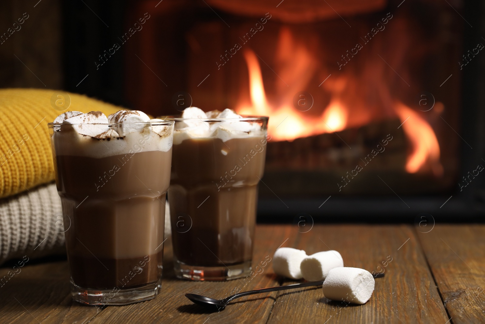 Photo of Glasses with hot cocoa, marshmallows and sweaters on wooden table near fireplace, space for text