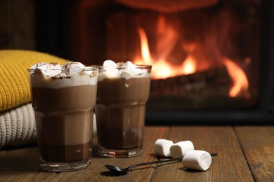 Photo of Glasses with hot cocoa, marshmallows and sweaters on wooden table near fireplace, space for text