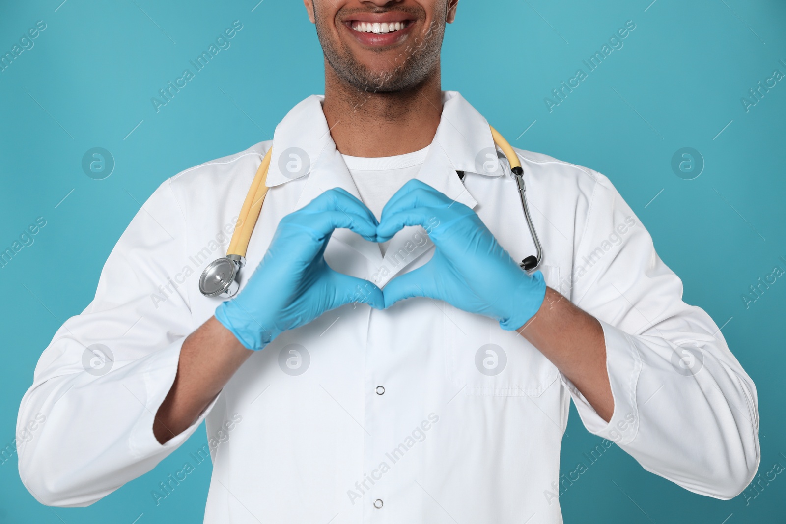 Photo of Doctor or medical assistant (male nurse) in uniform making heart with hands turquoise background, closeup