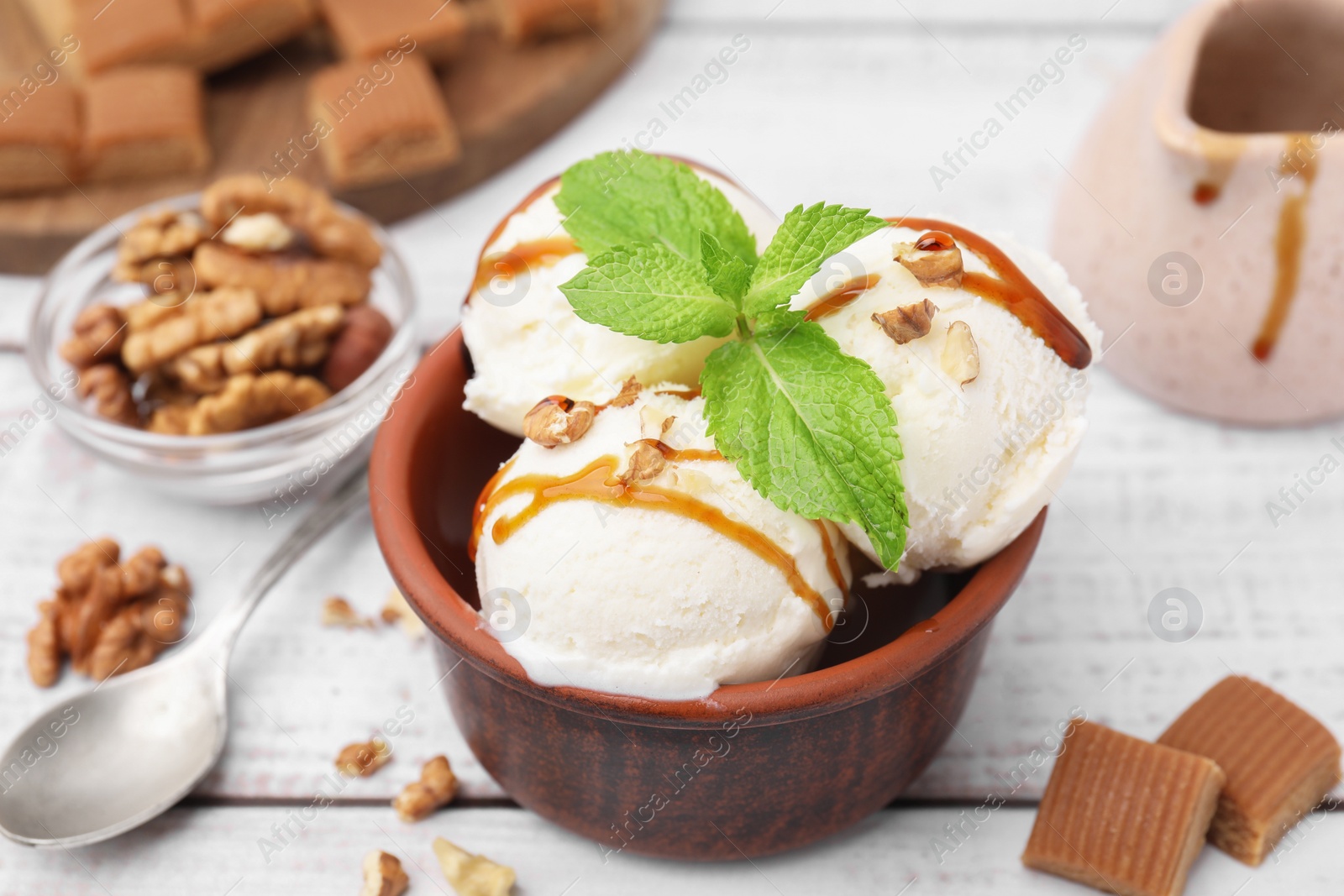 Photo of Bowl of tasty ice cream with caramel sauce, mint and nuts on white wooden table, closeup