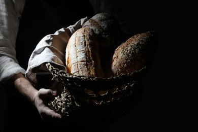 Photo of Man holding wicker basket with different types of bread on dark brown background, closeup