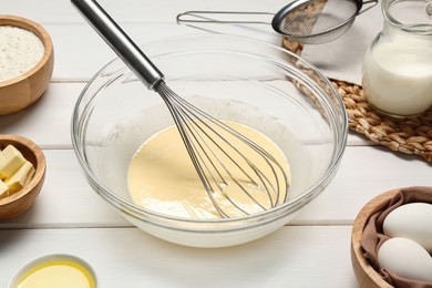 Photo of Glass bowl of crepe batter with whisk and ingredients on white wooden table