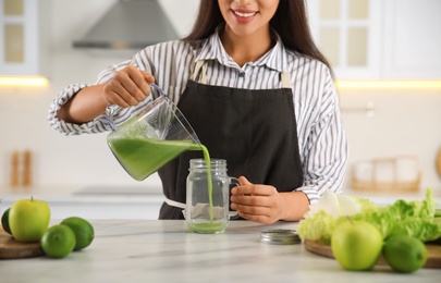Photo of Young woman pouring fresh green juice into mason jar at table in kitchen, closeup