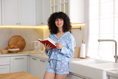 Beautiful young woman in stylish pyjama with book in kitchen