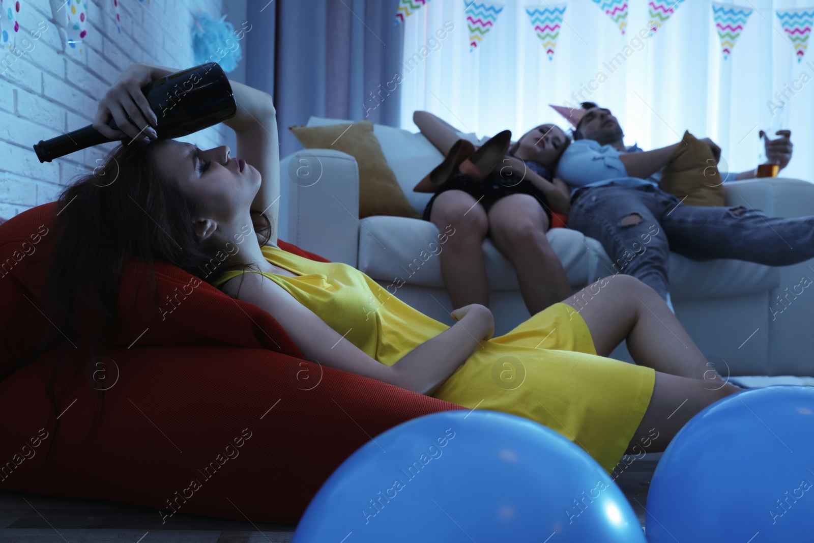 Photo of Young woman suffering from hangover and her friends sleeping on sofa in messy room after party