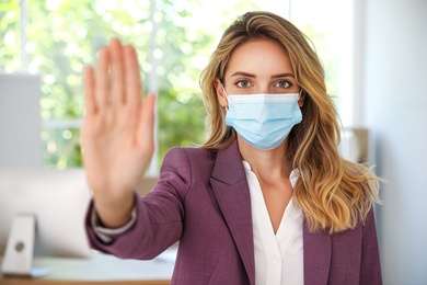 Woman in protective face mask showing stop gesture in office. Prevent spreading of coronavirus