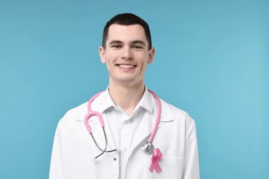 Portrait of smiling mammologist with pink ribbon and stethoscope on light blue background. Breast cancer awareness